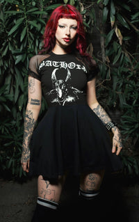 Person wearing a black dress with a graphic design, standing against a dark green leafy background.