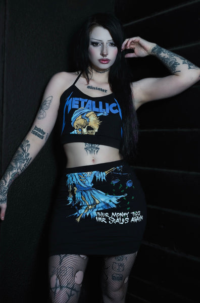 Person wearing a 'Metallica' tank top and skirt with tattoos, standing against a dark background.