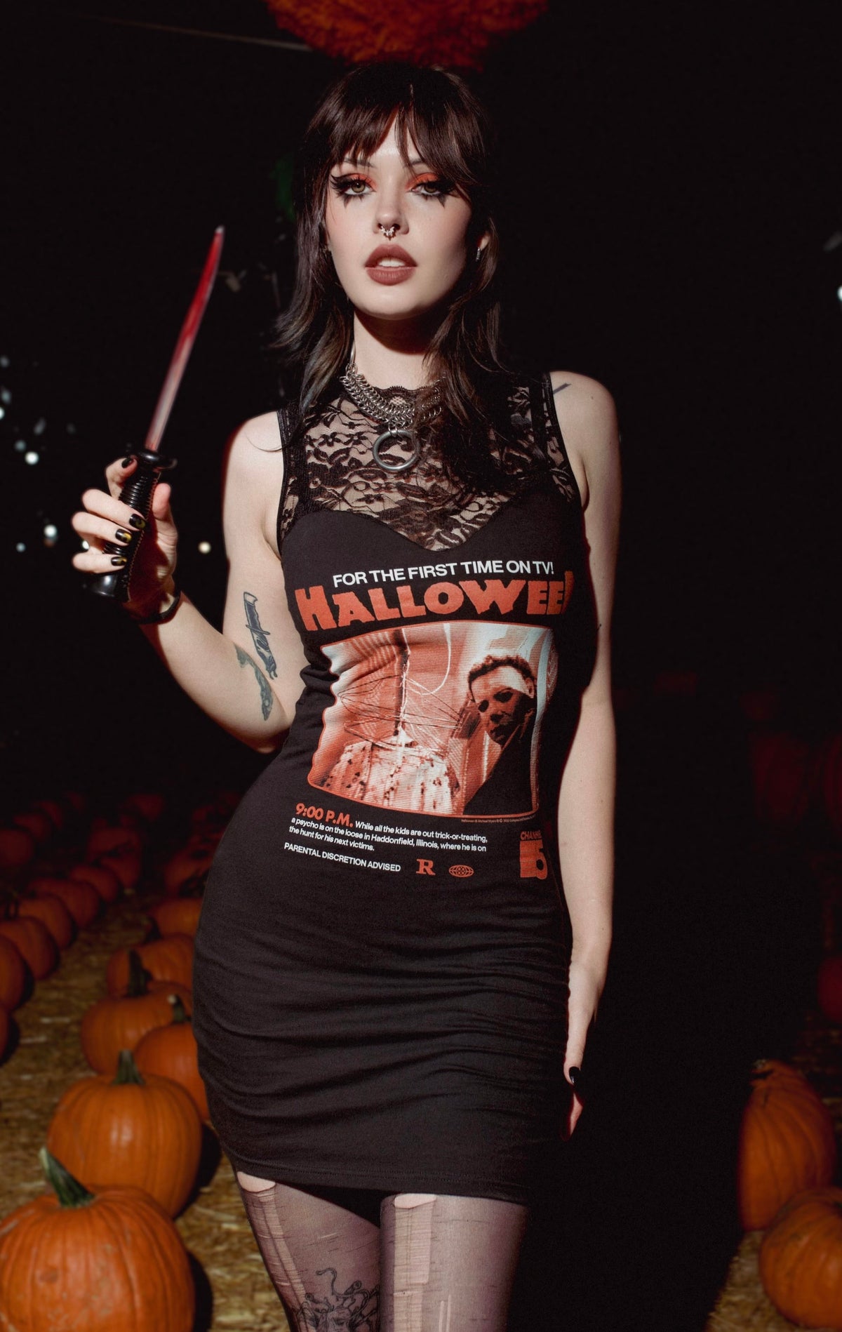 Woman in a Halloween-themed dress holding a knife among pumpkins at night.