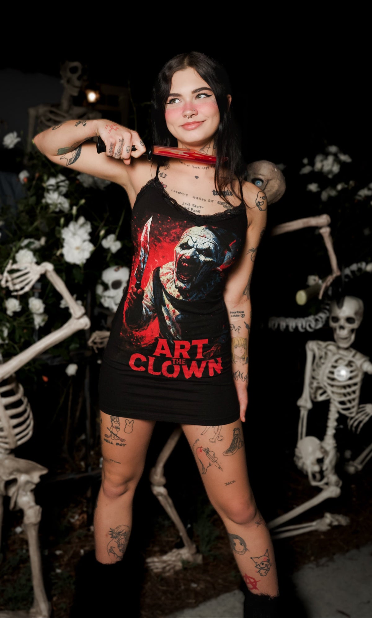 Person wearing a black dress with a graphic design and text, standing in front of skeletons and flowers at night.