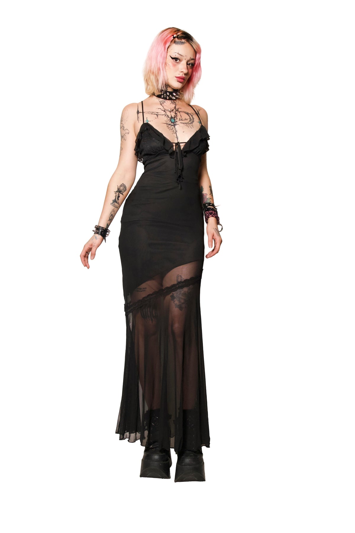 A model with pink hair wearing a black sheer lace slip dress with a corset-style bodice, sheer mesh skirt, and black platform boots, photographed against a solid white background.