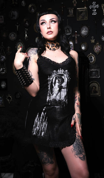 Person wearing a black dress with a graphic design, standing against a dark background with decorative elements.