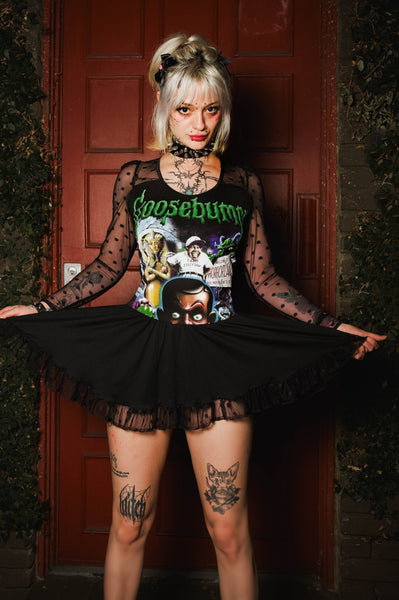 A woman stands in front of a dark red door wearing a black gothic-style costume top with sheer polka-dot sleeves, a graphic print featuring the word 'Coasebum' in green, and a black ruffled tutu skirt.