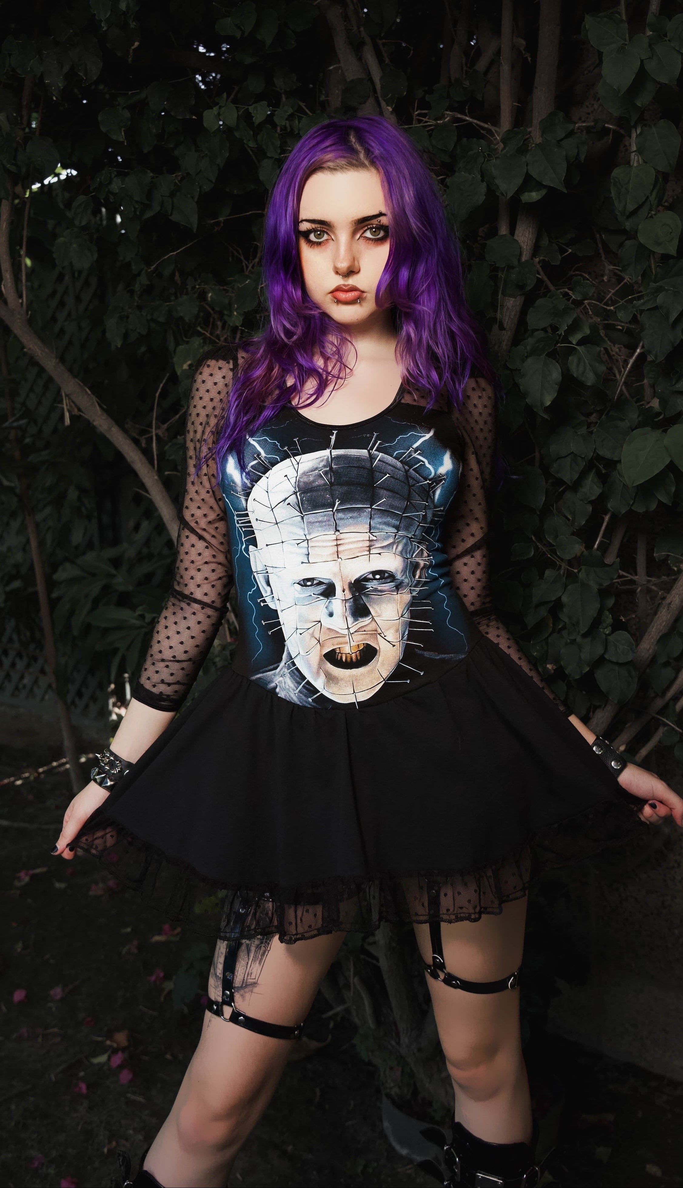 A person with bright purple hair stands outdoors in front of dark foliage, wearing a black gothic outfit with a graphic print on the front featuring a pale skull-like face, sheer black mesh sleeves, a black skirt, and leg straps.