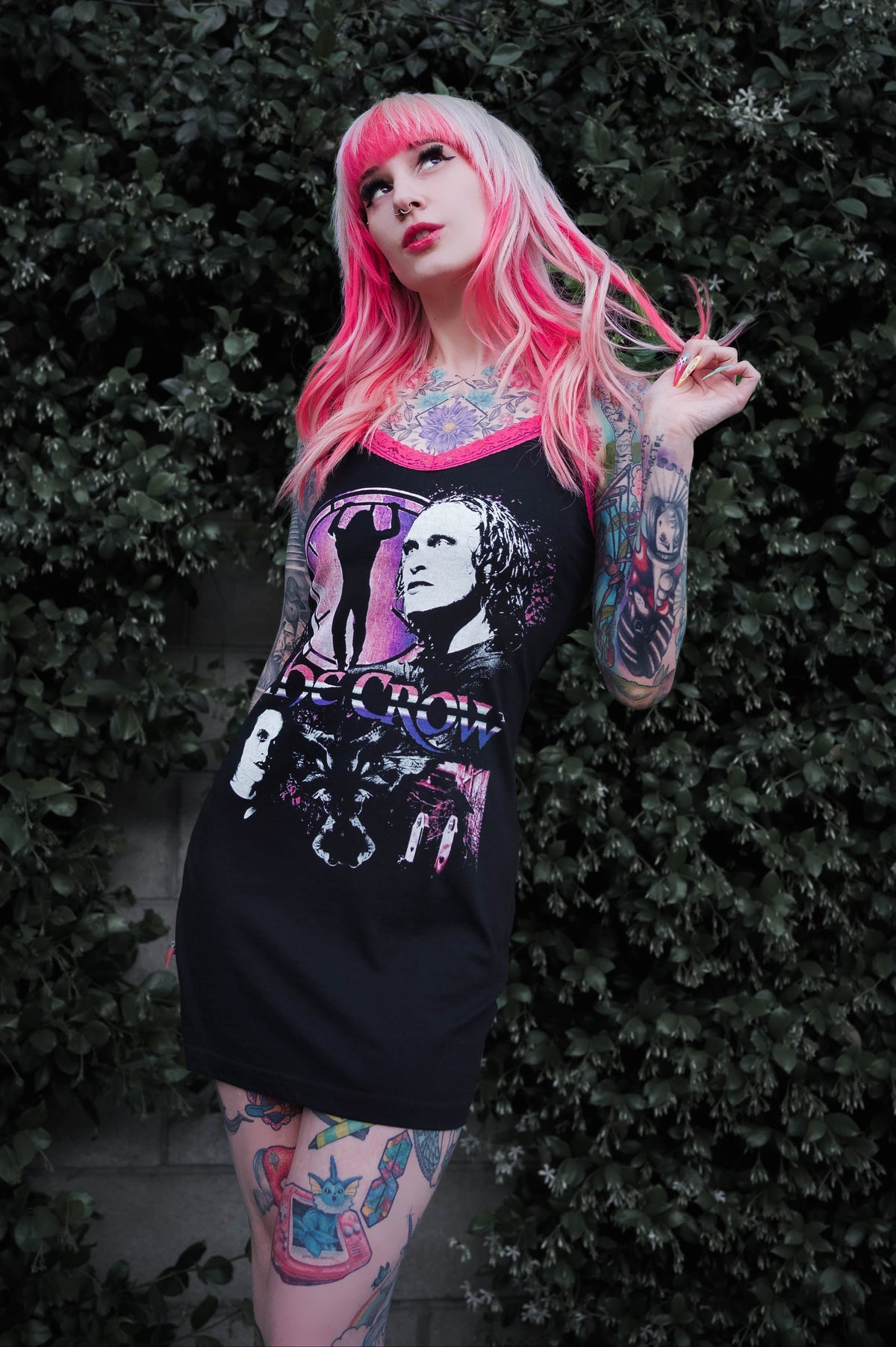 Person with pink hair and tattoos wearing a black dress with a graphic design against a green bush background
