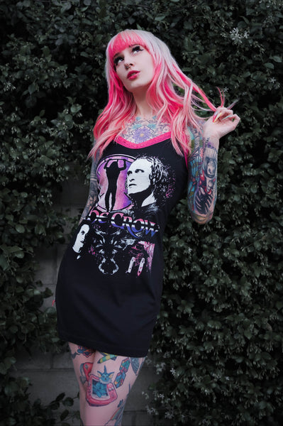 Person with pink hair and tattoos wearing a black dress with a graphic design against a green bush background