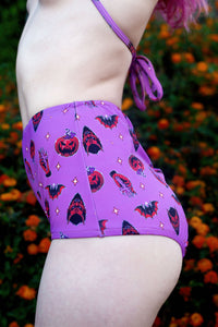 purple halter thin strap bikini top and high waist bikini bottom. Features black and orange images of pumpkins, bats, devil and hand holding cross