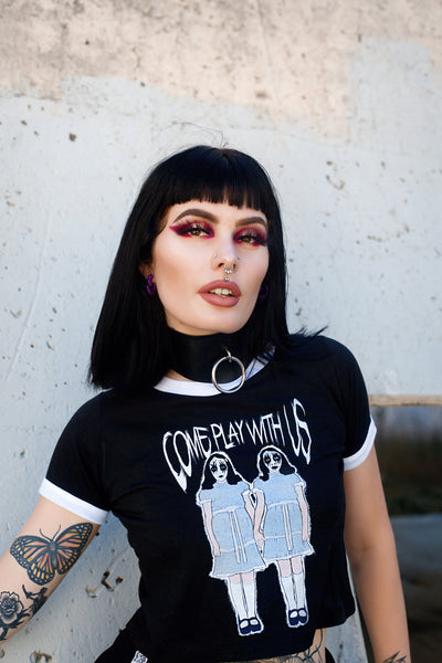 Black ringer tee with white trim. Features image of two girls holding hands with white and black face makeup wearing identical baby blue and white dresses with black shoes and white knee high stockings. Inscription reads "COME PLAY WITH US" in white.
