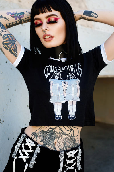 Black ringer tee with white trim. Features image of two girls holding hands with white and black face makeup wearing identical baby blue and white dresses with black shoes and white knee high stockings. Inscription reads "COME PLAY WITH US" in white.