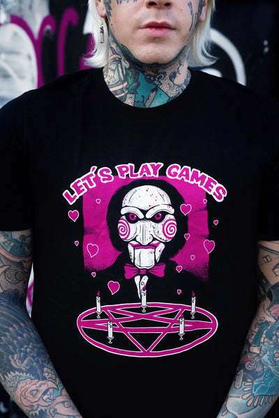 Black short sleeve t-shirt. Features face of Jigsaw from the movie Saw above a pink pentagram with white candles. Pink background and pink hearts around Jigsaw. Inscription reads "LET'S PLAY GAMES" in pink letters with white outline.