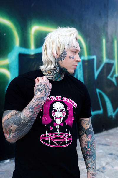 Black short sleeve t-shirt. Features face of Jigsaw from the movie Saw above a pink pentagram with white candles. Pink background and pink hearts around Jigsaw. Inscription reads "LET'S PLAY GAMES" in pink letters with white outline.