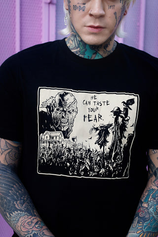 Black short sleeve t-shirt. Features black and white image of creeper from movie Jeepers Creepers face over house and cornfield with scarecrows and crows. Inscription reads "HE CAN TASTE YOUR FEAR" in black.