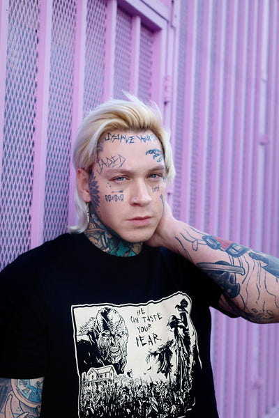 Black short sleeve t-shirt. Features black and white image of creeper from movie Jeepers Creepers face over house and cornfield with scarecrows and crows. Inscription reads "HE CAN TASTE YOUR FEAR" in black.