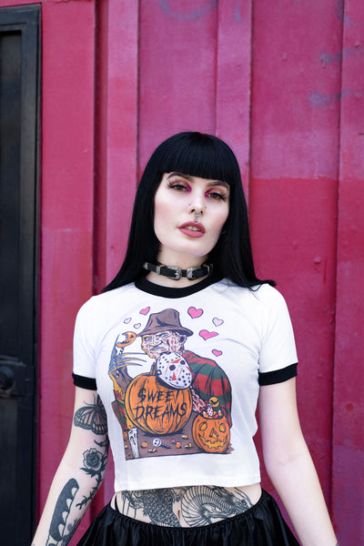 White ringer tee with black trim. Print is Freddy Krueger eating lollipop and has pumpkins and candy in front of him. On the pumpkins is a Jason hockey mask and pink hearts are behind Freddy. Center pumpkin says "Sweet Dreams" in black font
