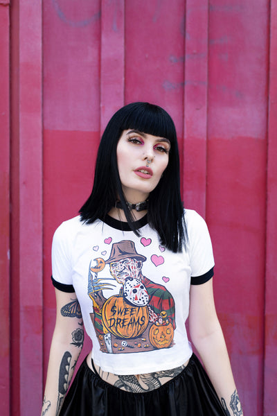 White ringer tee with black trim. Print is Freddy Krueger eating lollipop and has pumpkins and candy in front of him. On the pumpkins is a Jason hockey mask and pink hearts are behind Freddy. Center pumpkin says "Sweet Dreams" in black font