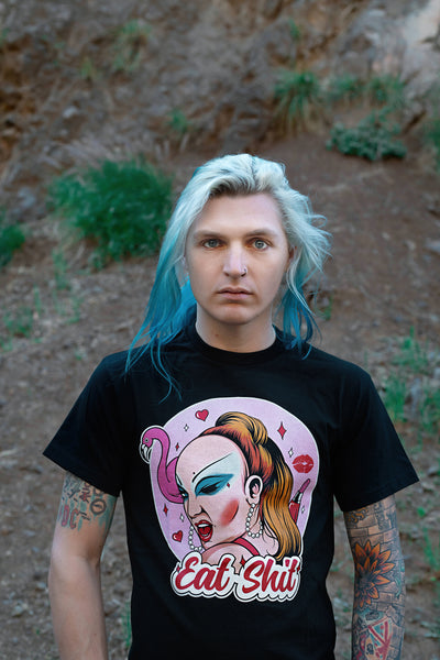 Black short sleeve t-shirt. Features woman with makeup winking her eye. Lipstick, hearts, and flamingo behind her with a pink background. Inscription reads "Eat Shit" written in pink font.