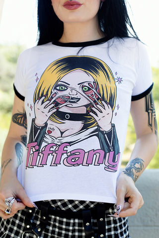 White ringer tee with black trim. Features Tiffany from the movie Bride of Chucky wearing black jacket and choker with her face melting. Black "Chucky" and red heart tattoo on her breast and pink sparkles in background of image. Inscription reads "Tiffany" in pink.
