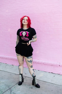 Black ringer tee with pink and black stripped trim. Features face of Jigsaw from the movie Saw above a pink pentagram with white candles. Pink background and pink hearts around Jigsaw. Inscription reads "LET'S PLAY GAMES" in pink letters with white outline.
