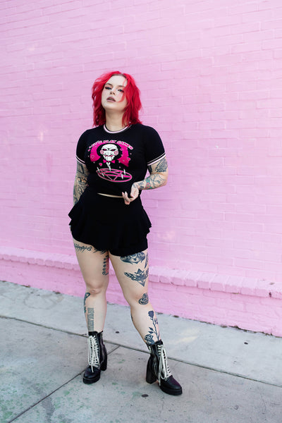 Black ringer tee with pink and black stripped trim. Features face of Jigsaw from the movie Saw above a pink pentagram with white candles. Pink background and pink hearts around Jigsaw. Inscription reads "LET'S PLAY GAMES" in pink letters with white outline.