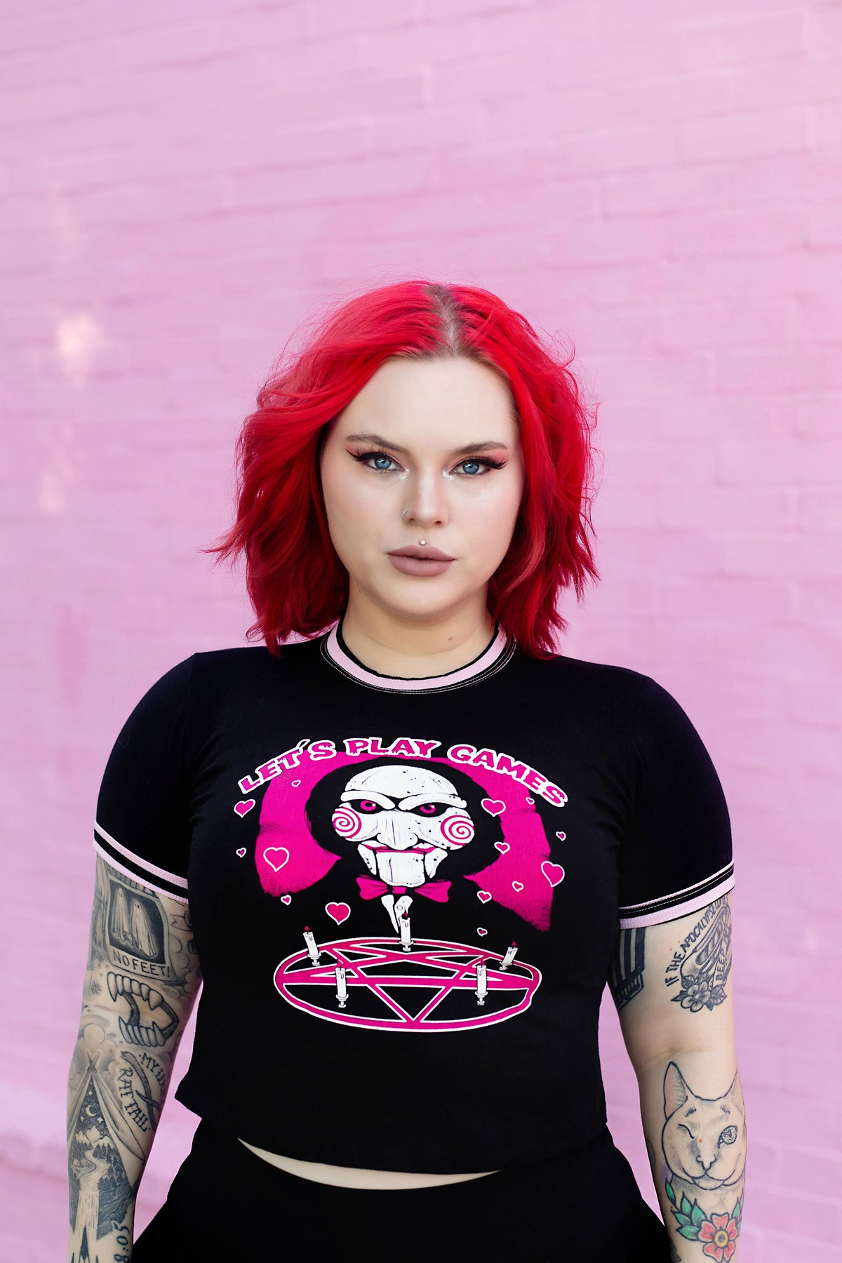 Black ringer tee with pink and black stripped trim. Features face of Jigsaw from the movie Saw above a pink pentagram with white candles. Pink background and pink hearts around Jigsaw. Inscription reads "LET'S PLAY GAMES" in pink letters with white outline.