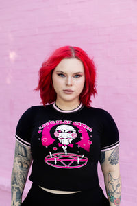 Black ringer tee with pink and black stripped trim. Features face of Jigsaw from the movie Saw above a pink pentagram with white candles. Pink background and pink hearts around Jigsaw. Inscription reads "LET'S PLAY GAMES" in pink letters with white outline.