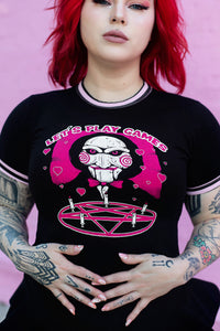 Black ringer tee with pink and black stripped trim. Features face of Jigsaw from the movie Saw above a pink pentagram with white candles. Pink background and pink hearts around Jigsaw. Inscription reads "LET'S PLAY GAMES" in pink letters with white outline.