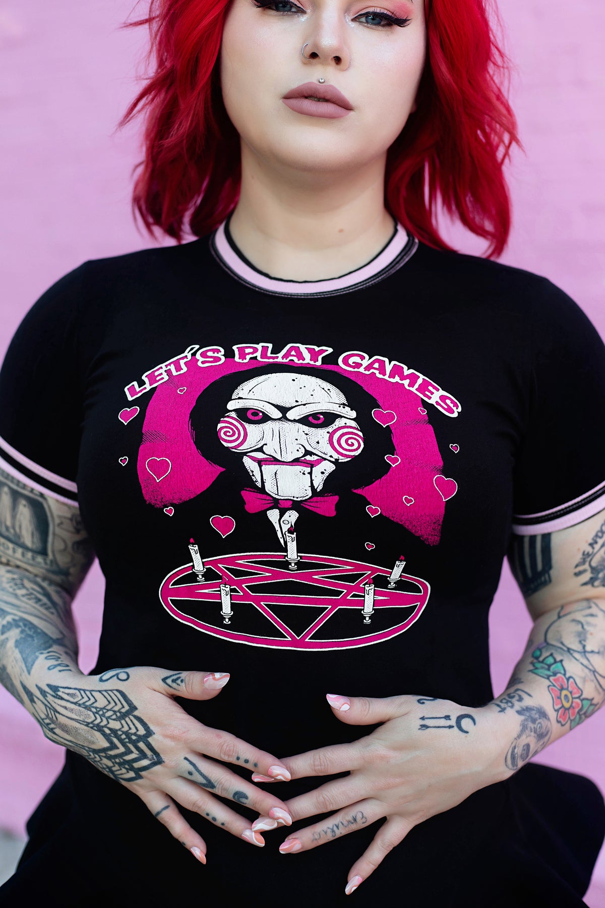 Black ringer tee with pink and black stripped trim. Features face of Jigsaw from the movie Saw above a pink pentagram with white candles. Pink background and pink hearts around Jigsaw. Inscription reads "LET'S PLAY GAMES" in pink letters with white outline.