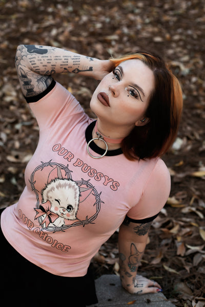 Baby pink ringer tee with black trim. Features image of a white cat wearing a pink bow winking inside a bob wire heart. Inscriptions reads "OUR PUSSY OUR CHOICE" in pink.