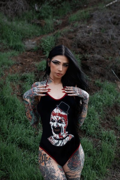 Black body suit with thin red trim neckline and thin red straps to tie around neck. Features clown from the movie Terrifier wearing a black top hat with red blood splattered on face.