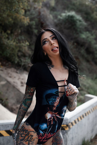 Black short sleeve bodysuit with black lace up V neck. Features Freddy Krueger holding knife glove up to woman's face with houses below. Inscription reads "A Nightmare on Elm Street" in red.