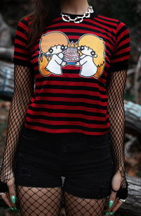 Black and red stripped ringer tee with black trim. Features image of cartoon boy and girl wearing white dresses with black makeup on eyes and black upside down cross on foreheads. They are holding a white cake with pink frosting and pink 666 candles lit on cake. 