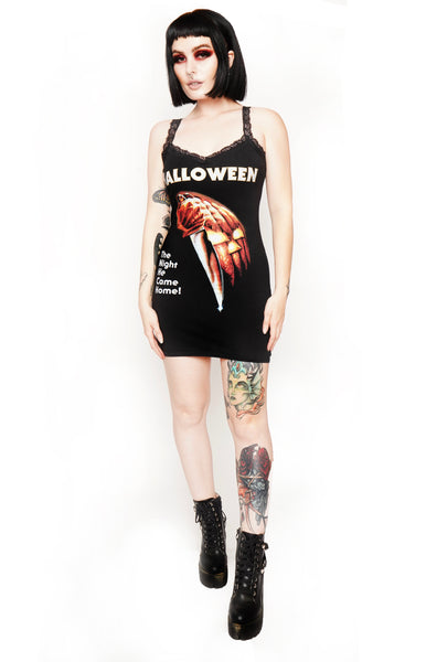Black mini dress with thin lace straps and black lace trim neckline. Print features jack o lantern with knife. Inscription reads "Halloween" and "The Night He Comes Home" in white font