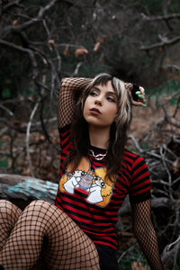 Black and red stripped ringer tee with black trim. Features image of cartoon boy and girl wearing white dresses with black makeup on eyes and black upside down cross on foreheads. They are holding a white cake with pink frosting and pink 666 candles lit on cake. 