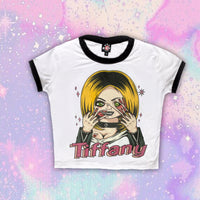White ringer tee with black trim. Features Tiffany from the movie Bride of Chucky wearing black jacket and choker with her face melting. Black "Chucky" and red heart tattoo on her breast and pink sparkles in background of image. Inscription reads "Tiffany" in pink.
