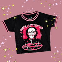 Black ringer tee with pink and black stripped trim. Features face of Jigsaw from the movie Saw above a pink pentagram with white candles. Pink background and pink hearts around Jigsaw. Inscription reads "LET'S PLAY GAMES" in pink letters with white outline.