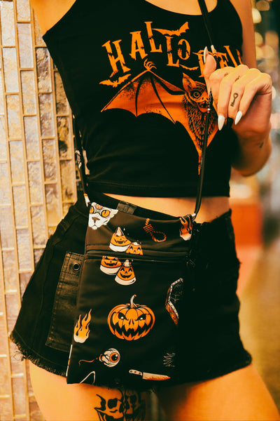 Black mini concert style purse with thin straps and zipper. Print is Halloween pattern containing candy corn, flames, cat, coffins, pumpkins, poison bottle, noose, and crow in black white and orange.