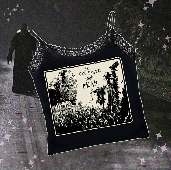 Black thin strap tank top with black lace trim neckline. Features black and white image of creeper from movie Jeepers Creepers face over house and cornfield with scarecrows and crows. Inscription reads "HE CAN TASTE YOUR FEAR" in black.