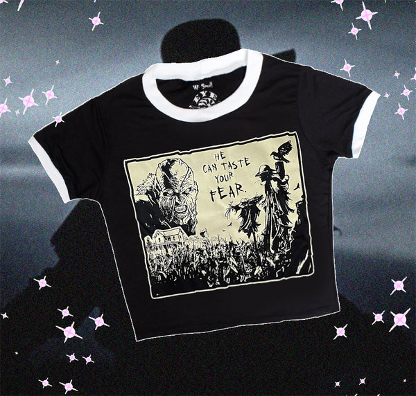 Black ringer tee with white trim. Features black and white image of creeper from movie Jeepers Creepers face over house and cornfield with scarecrows and crows. Inscription reads "HE CAN TASTE YOUR FEAR" in black.