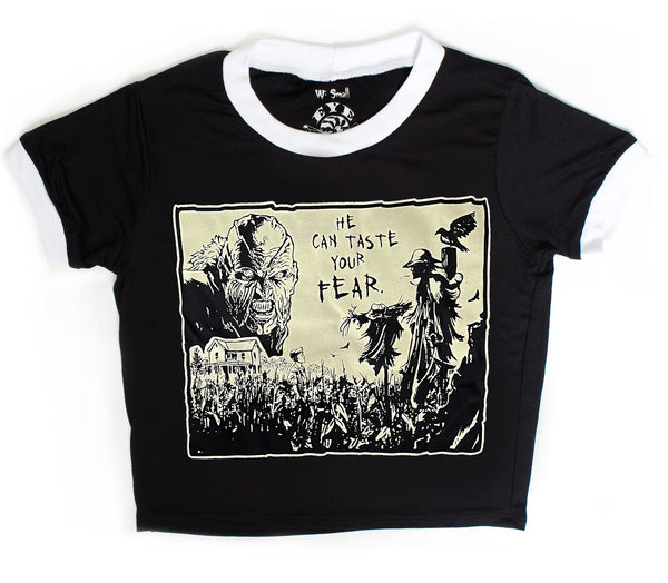 Black ringer tee with white trim. Features black and white image of creeper from movie Jeepers Creepers face over house and cornfield with scarecrows and crows. Inscription reads "HE CAN TASTE YOUR FEAR" in black.