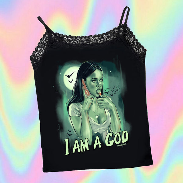 Black thin strap tank top with black lace trim neckline. Features green print image of Jennifer from the movie Jennifer's body holding a cell phone to her ear and a lighter to her tongue. Full moon and bats in background. Inscription reads "I AM A GOD" in green.