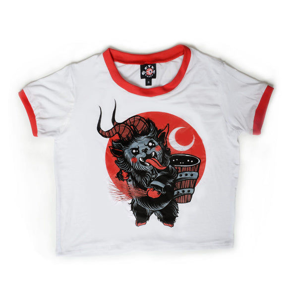 White ringer tee with red trim. Features animated Krampus sticking his tongue out while holding sticks in hand and wood pail on back. Red background with white crescent moon.