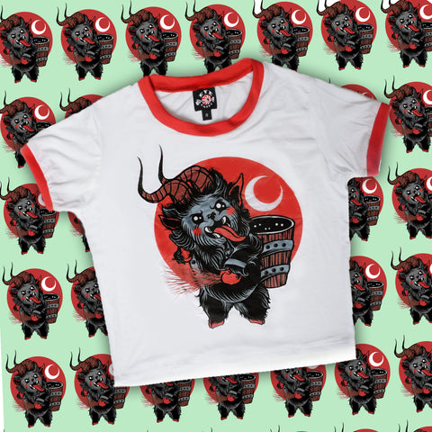 White ringer tee with red trim. Features animated Krampus sticking his tongue out while holding sticks in hand and wood pail on back. Red background with white crescent moon.