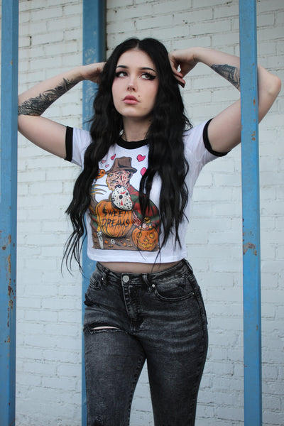 White ringer tee with black trim. Print is Freddy Krueger eating lollipop and has pumpkins and candy in front of him. On the pumpkins is a Jason hockey mask and pink hearts are behind Freddy. Center pumpkin says "Sweet Dreams" in black font
