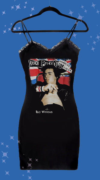 Black thin strap mini dress with black lace trim neckline. Features image of singer Sid Vicious sitting back on ref chair. Inscription reads "Sex Pistols" and "ANARCHY IN THE UK" in black. Inscription in white reads "Sid Vicious". Image of U.K. flag in background.