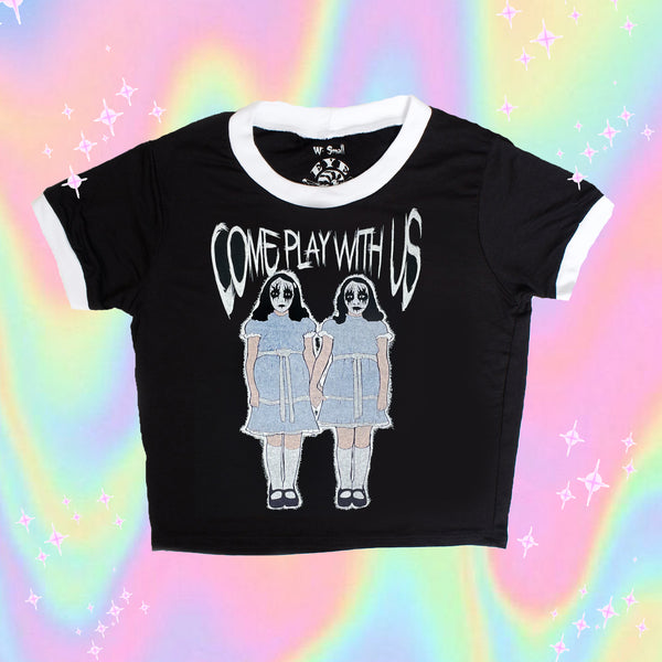 Black ringer tee with white trim. Features image of two girls holding hands with white and black face makeup wearing identical baby blue and white dresses with black shoes and white knee high stockings. Inscription reads "COME PLAY WITH US" in white.