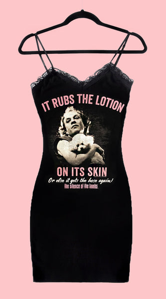 Black thin strap mini dress with black lace trim neckline. Features white print image of Ted Levine from the movie The Silence of The Lambs holding his white dog. Inscription reads "IT RUBS THE LOTION ON ITS SKIN OR ELSE IT GETS THE HOSE AGAIN!" in pink and white and "THE SILENCE OF THE LAMBS" in pink.